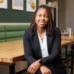 SheilaPhicil Sheila Phicil wearing black suit, white blouse. Sitting in front of a table